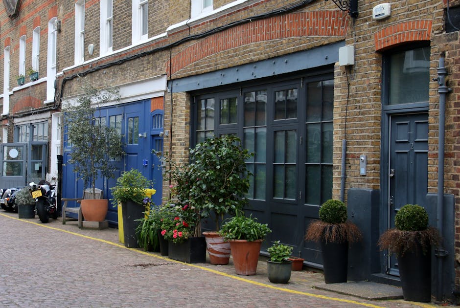 A close-up view of a red brick residential building on Mortlake High Street, showing two white-framed sash windows with closed blinds on a ground-floor level. To the right of the windows, there is a d
